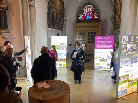 Besucher betrachten eine LED-Ausstellung mit leuchtenden Infodisplays in der dunklen Kirche St. Agatha in Mettingen – realisiert von der Wrocklage Werbewerkstatt.