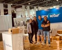 Gruppenfoto vor dem frisch aufgebauten, noch nicht dekorierten Messestand mit sichtbaren Kartons und Equipment