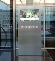 Hochwertige Edelstahlstele mit beleuchtetem Schriftzug „PHOENIX“ und integriertem Touchdisplay, auf dem ein digitales Menü angezeigt wird. Die Stele steht im Eingangsbereich eines modernen Gebäudes mit Glasfassade.