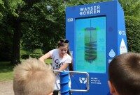 Kinder bedienen blaue Station zum Wasserbohren bei interaktiver Ausstellung im Park.