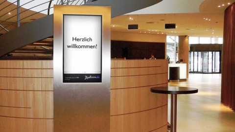 : Digitale Stele mit „Herzlich willkommen!“-Bildschirm im modernen Empfangsbereich des Radisson Blu Hotels Hamburg Airport.