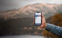 Hand hält Smartphone mit geöffneter App zur Zeiterfassung vor unscharfem Bergpanorama im Hintergrund.