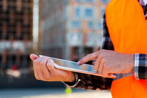 Person mit orangefarbener Warnweste bedient Tablet auf Baustelle zur Materialverfolgung.