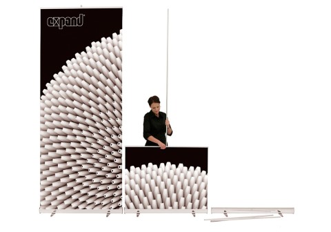 Expand Roll-Up Banner System – flexibles Werbedisplay mit einfachem Aufbau Frau beim Aufbau eines Expand Roll-Up Banners mit modernem grafischen Design aus weißen Rollen auf schwarzem Hintergrund