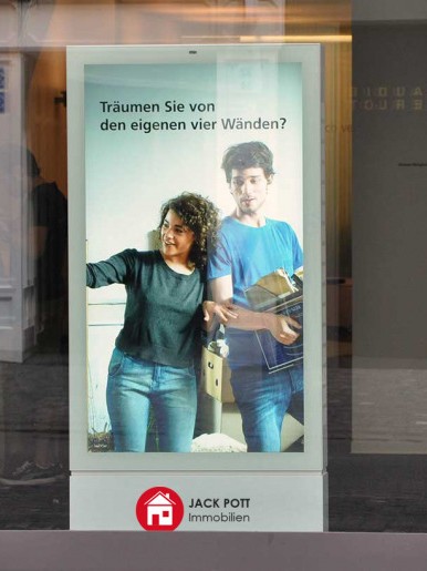 Digitale Werbestele in einer Fußgängerzone zeigt ein Paar mit Umzugskartons und dem Text „Träumen Sie von den eigenen vier Wänden?“. Unten das Logo von Jack Pott Immobilien. Beispiel für digitale Immobilienwerbung im urbanen Raum.