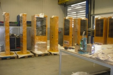 Lagerraum mit mehreren orange lackierten, in Folie verpackten Kiosksystemen auf Paletten.