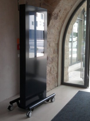 Ein schwarzer Stele Rollwagen mit einem Display auf Rollen neben einer gläsernen Tür in einem Gebäude.