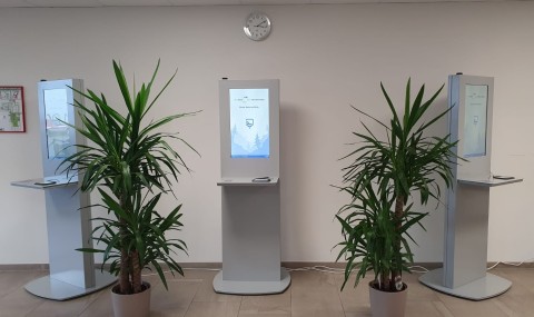 Drei Stehterminals mit digitalen Bildschirmen, umgeben von Pflanzen in einem Flur