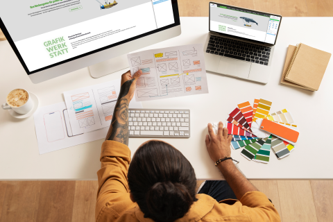 Grafikdesigner bei der Arbeit mit Farbfächern und Webdesign-Entwürfen Designer am Schreibtisch mit Farbmustern, Skizzen und Computer beim Erstellen von Webdesign-Layouts